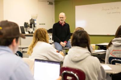 Instructor seated at the front of a classroom while students face him.